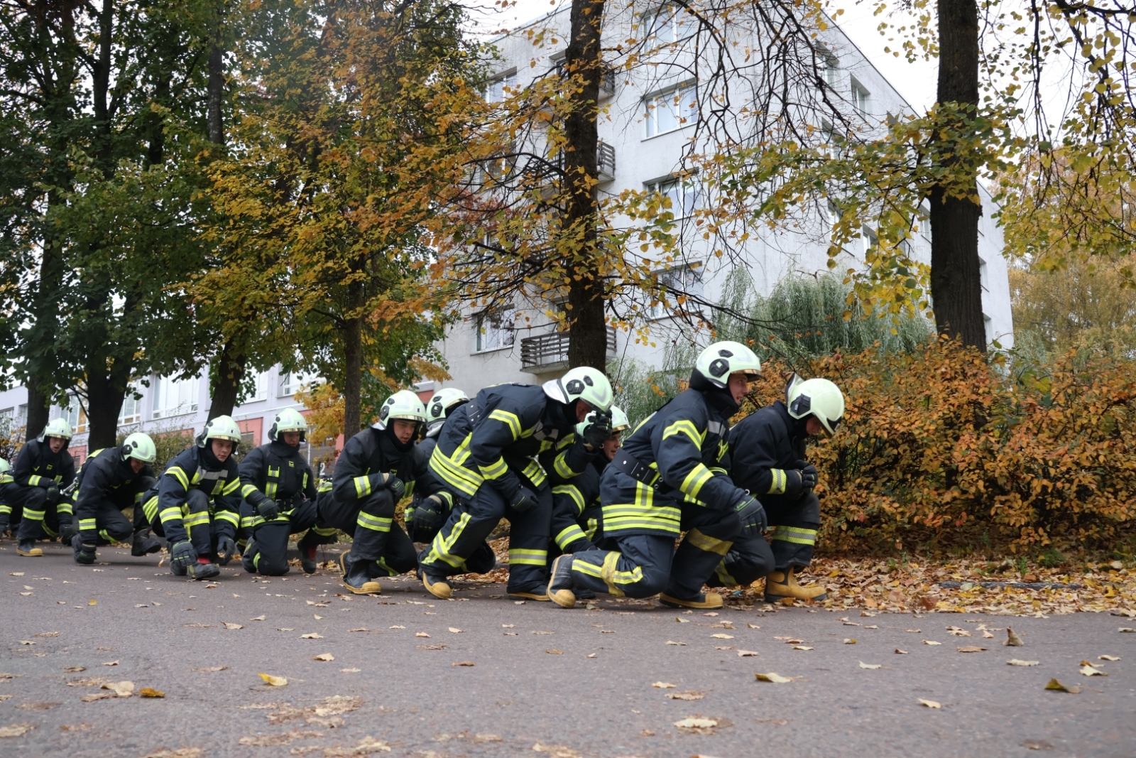 Koledžas kadeti, ģērbušies aizsargtērpos, pārvietojas "zosu gājienā". Fonā koki un balta ēka.