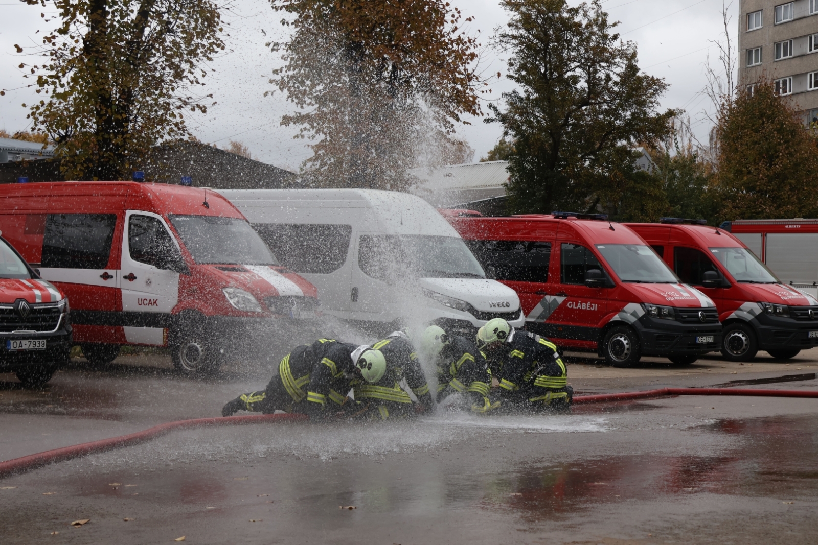 Koledžas kadeti, ģērbušies aizsargtērpos, noliekušies pie ūdens šļūtenes, no kuras plūst ūdens. Fonā VUGD automašīnas.