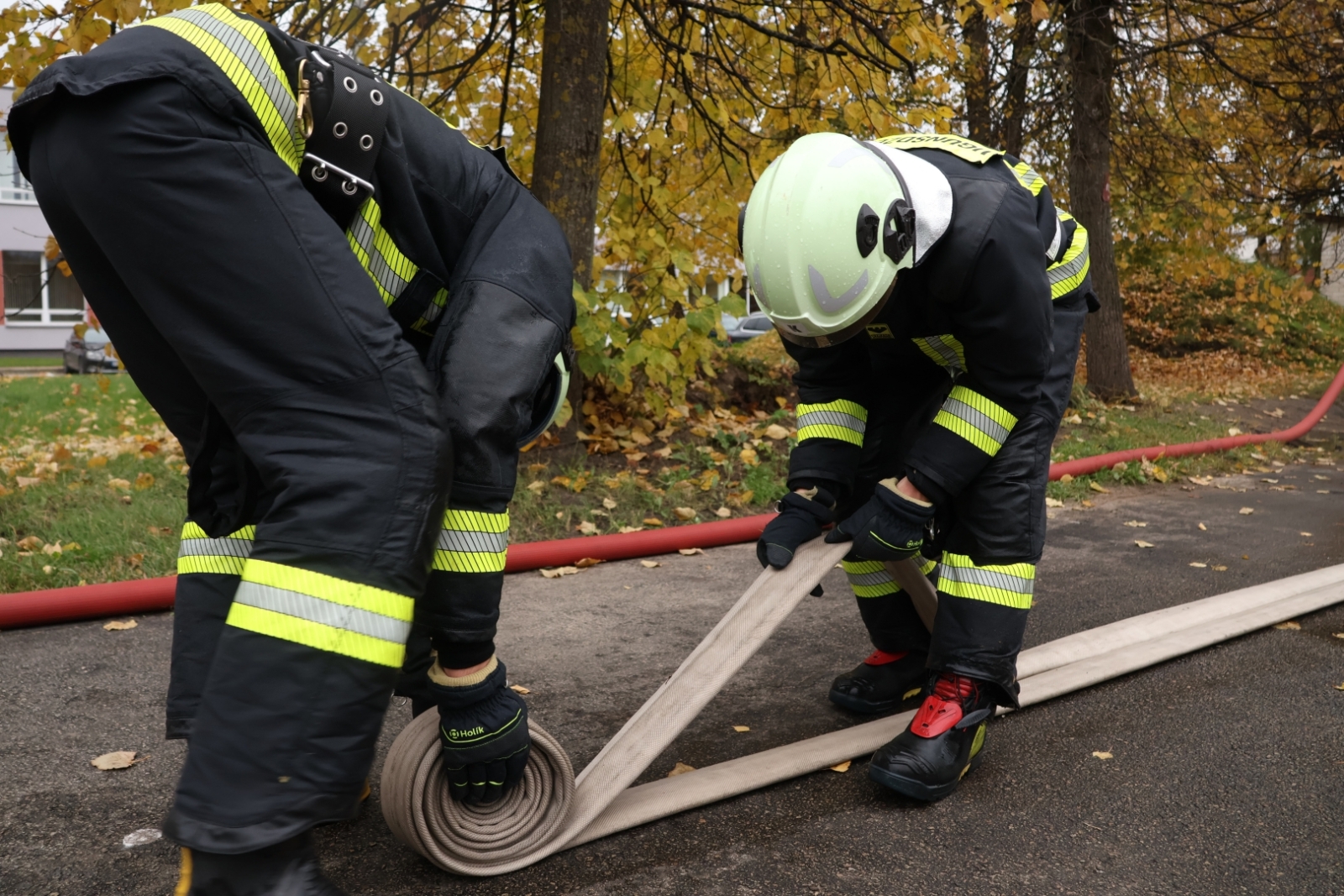 Divi koledžas kadeti, ģērbušies aizsargtērpos, ritina ugunsdzēsības šļūteni.
