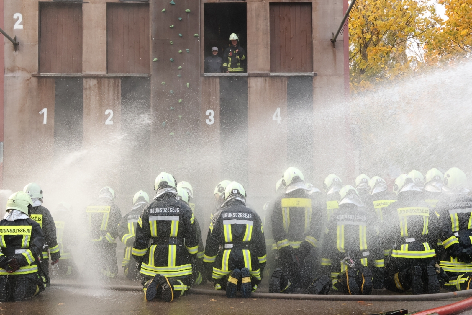 Koledžas kadeti no aizmugures - ģērbušies aizsargtērpos, notupušies ceļos. Kadeti tiek aplaistīti ar ūdeni. Fonā ēka, vienā no ēkas logiem redzami divi VUGD nodarbinātie.