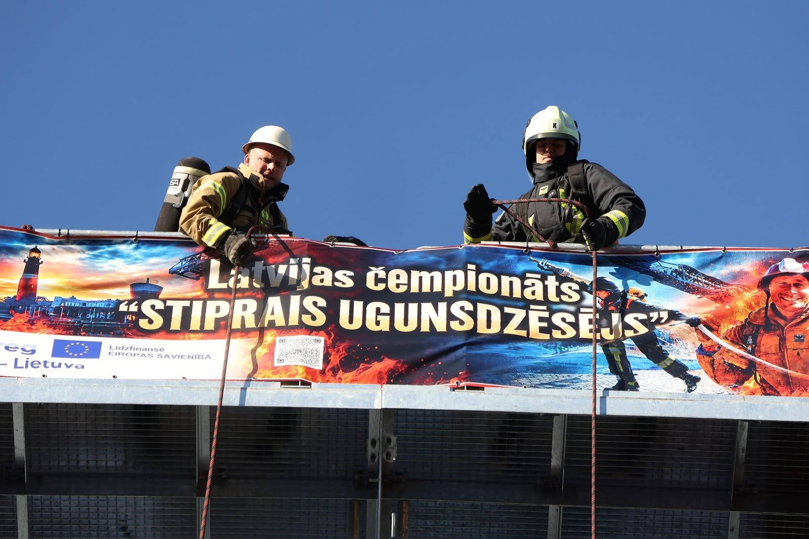 Uz sastatnēm stāv divi VUGD nodarbinātie aizsargtērpos. Uz stastatnēm plakāts, uz kura uzraksts "Latvijas cempionāts Stiprais ugunsdzēsējs"