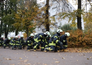 Koledžas kadeti, ģērbušies aizsargtērpos, pārvietojas "zosu gājienā". Fonā koki un balta ēka.