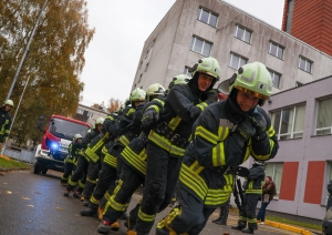 koledžas kadati, ģērbušies aizsargtērpos, velt ugunsdzēsības automašīnu. Labajā pusē fonā ēka, kreisajā pusē redzami koki.