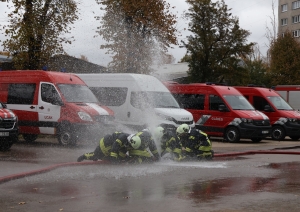 Koledžas kadeti, ģērbušies aizsargtērpos, noliekušies pie ūdens šļūtenes, no kuras plūst ūdens. Fonā VUGD automašīnas.