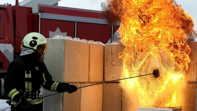 Tiek veikts paraugdemonstējums negadījumam, kas saistīts ar eļļas degšanu. Kreisajā pusē stāv ugundzēsējs glābējs. Labajā pusē plīts, uz kuras aizdedzies katls.