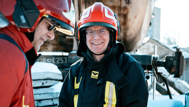 Valsts prezidents Edgars Rinkēvičs stāv ārā sniegotā teritorijā, smaida. Viņam uzvilkts ugunsdzēsēja glābēja formas tērps.