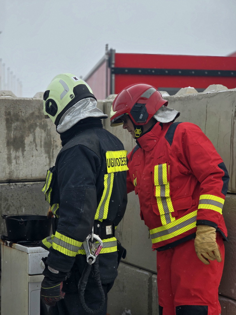 Divi ugunsdzēsēji glābēji gatavojas veikt paraugdemonstējumu, kas saistīts ar eļļas degšanu. Ugunsdzēsēji glābēji stāv ar mugurpusi, ar skatu uz plīti, uz kuras stāv katls.
