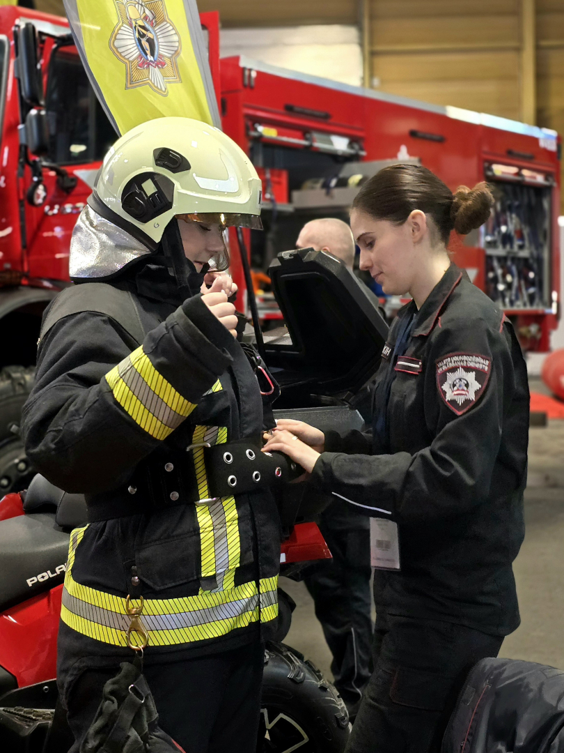 Ugunsdrošības un civilās aizsardzības koledžas kadete palīdz skolēnam piemērīt ugunsdzēsēja glābēja aizsargtērpu. 