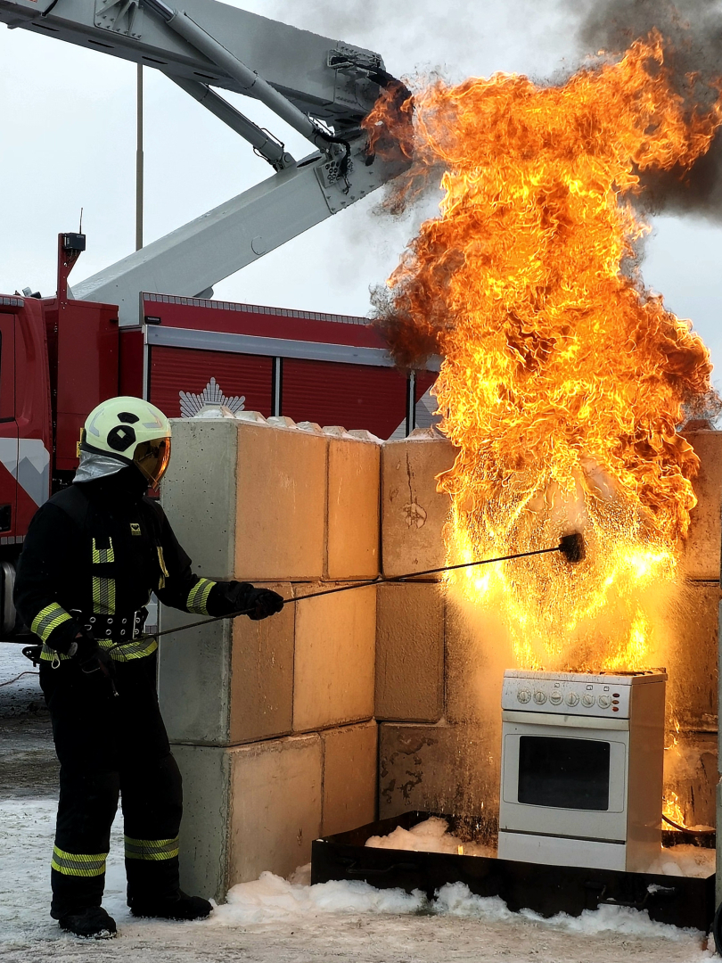 Tiek veikts paraugdemonstējums negadījumam, kas saistīts ar eļļas degšanu. Kreisajā pusē stāv ugundzēsējs glābējs. Labajā pusē plīts, uz kuras aizdedzies katls.