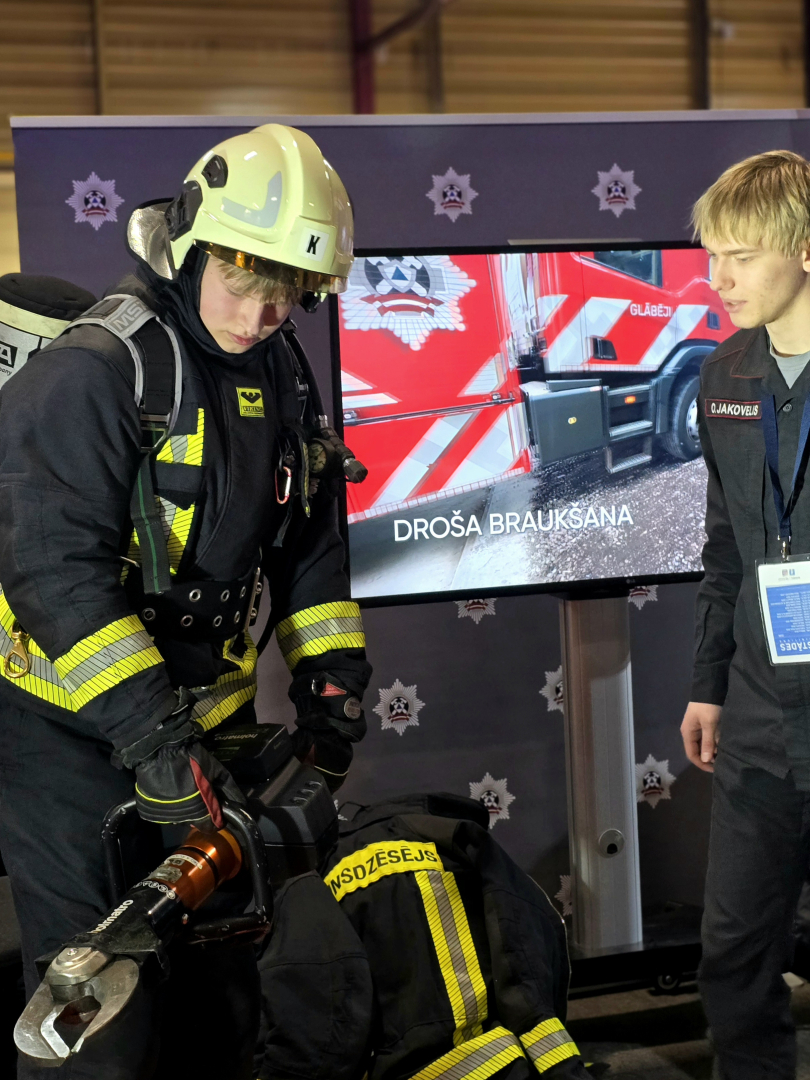 Skolēns uzvilcis ugunsdzēsēja glābēja aizsargtērpu. Viņam labajā pusē stāv Ugunsdrošības un civilās aizsardzības koledžas kadets.