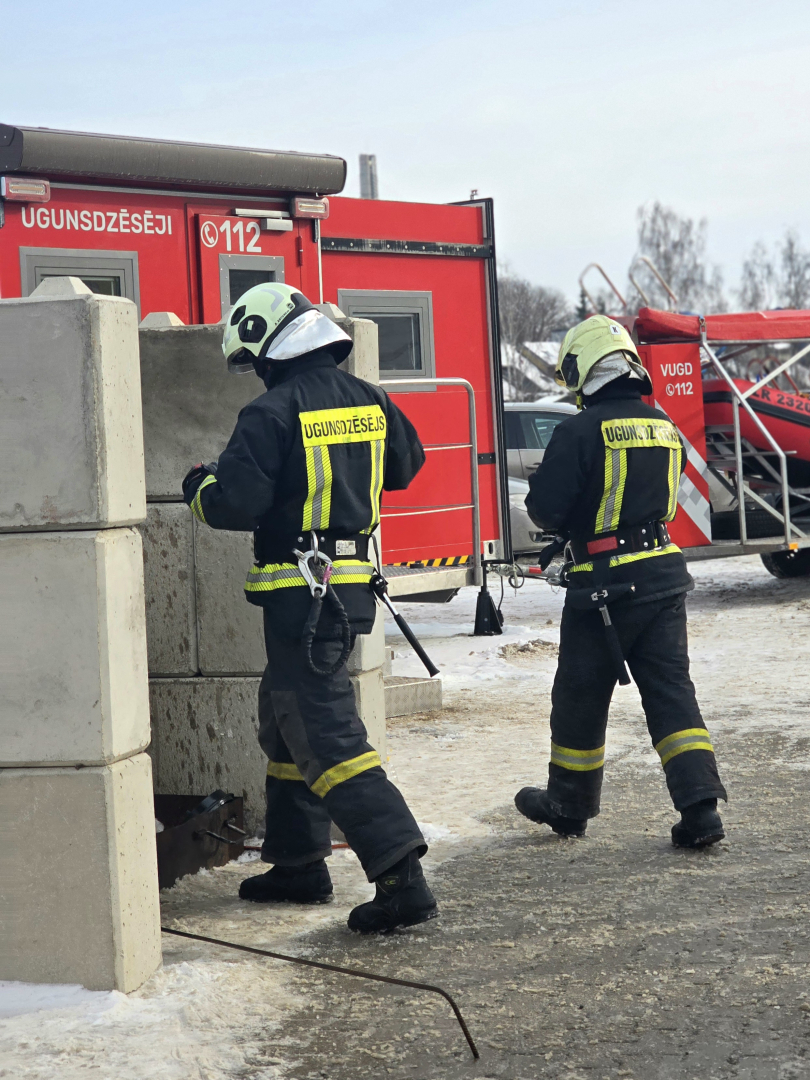 Divi ugunsdzēsēji glābēji no mugurpuses. Fonā Valsts ugunsdzēsības un glābšanas dienesta transportlīdzekļi. 