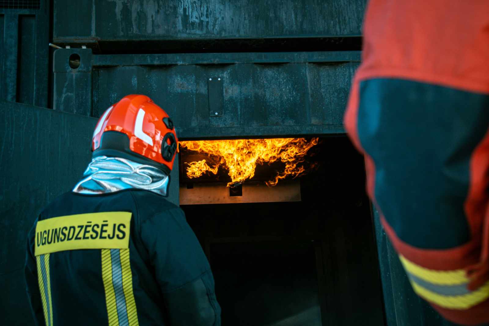 Ugunsdzēsējs glābējs stāv atvērtām ēkas durvīm. Ēkas iekšpusē redzamas liesmas.