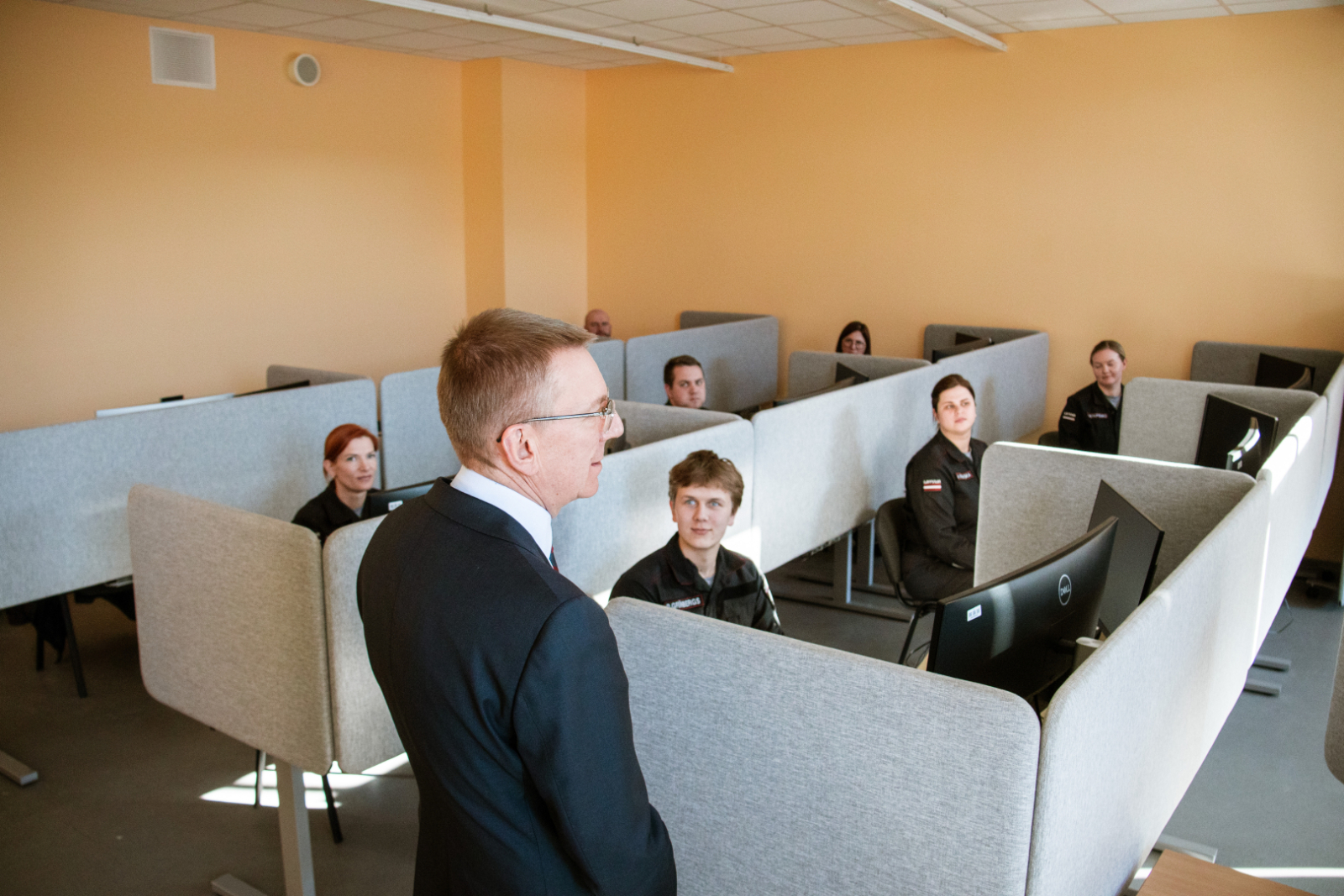 Priekšplānā ar mugurpusei nostājies valsts prezidents Edgars Rinkēvičs. Viņš pavērsies pret koledžas kadetiem, kuri sēž pie datoriem atsevišķās, ar paneļiem norobežotās vietās.